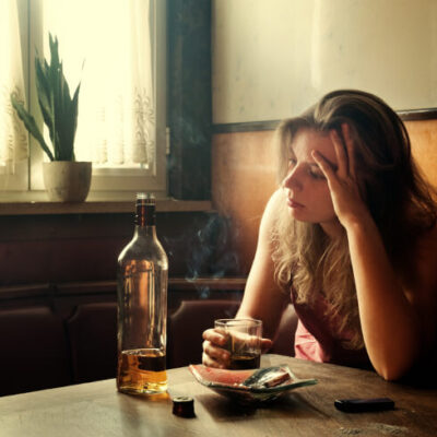 [Kobieta alkoholiczka siedząca przy stole, na którym jest alkohol i popielniczka – odnowa24h.pl]